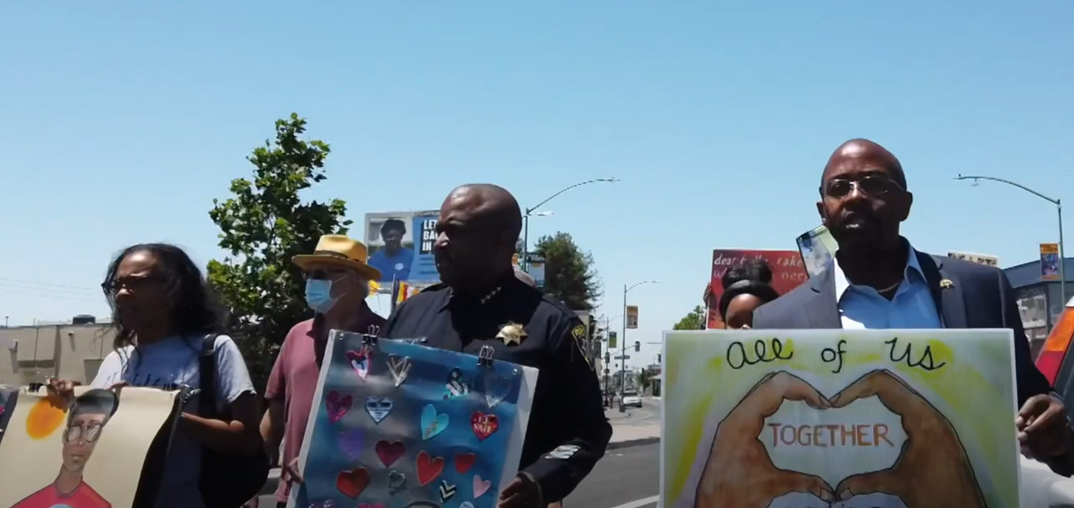 Still from Vision Quilt video of march with Loren Taylor on right holding a sign that says "All of us together"